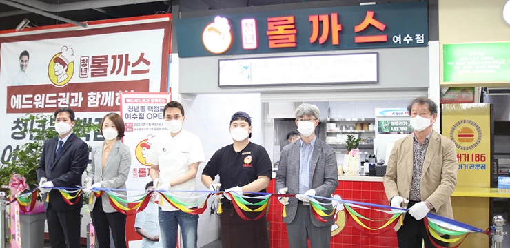 여수 중앙시장 청년몰(공식명칭 ‘꿈뜨락몰’)에 청년몰 핵점포 양성 지원사업으로 청년롤까스 여수점이 지난 9일 첫 영업을 시작했다. (에드워드 권(왼쪽에서 세 번째)과 청년 상인(왼쪽에서 네 번째), 여수시, 소상공인시장진흥공단, 청년상인육성재단, 중앙시장 상인회 관계자가 테이프 컷팅을 하고 있다.)