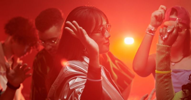 붉은 네온 빛으로 조명 나이트 클럽에서 그녀의 친구들 사이에서 움직이는 어두운 긴 머리를 가진 젊은 역동적인 여성 댄서의 측면 보기 / 셔터스톡