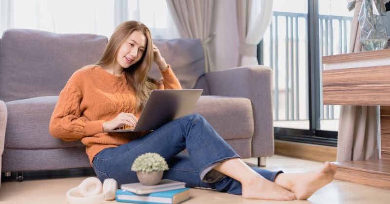 노트북을 사용 하 고 거실에서 소파 근처 바닥에 앉아 집에서 일 하 고 그녀의 여가 시간을 즐기는 웃는 젊은 아시아 여자 / 셔터스톡