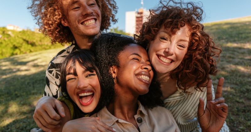 초록 풀과 나무들로 가득한 화창한 공원에서 함께 시간을 즐기고 있는 유쾌한 네 명의 젊은 친구들이 포옹하고 웃고 있습니다 / 셔터스톡