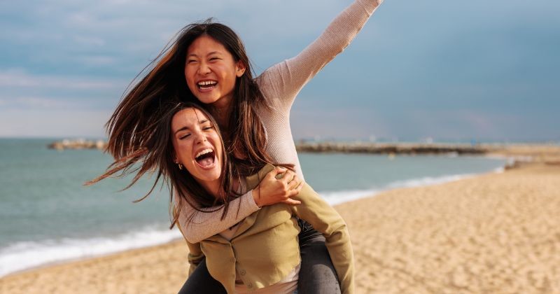 해변에서 빈티지 옷을 입고 서로 업어주며 활짝 웃고 있는 행복한 두 여사친 / 셔터스톡