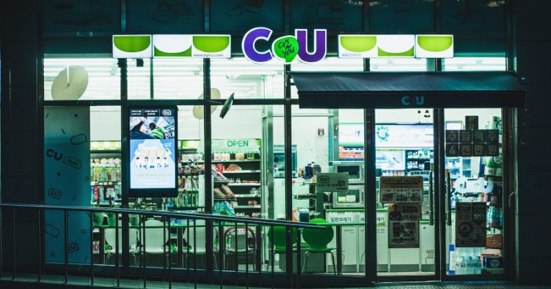 보라색과 초록색 로고가 밝게 빛나는 밤의 CU 편의점 외관으로, 환한 통유리창 너머로 진열된 상품들과 취식 공간이 들여다보이는 현대적인 한국 거리의 풍경입니다. / 셔터스톡