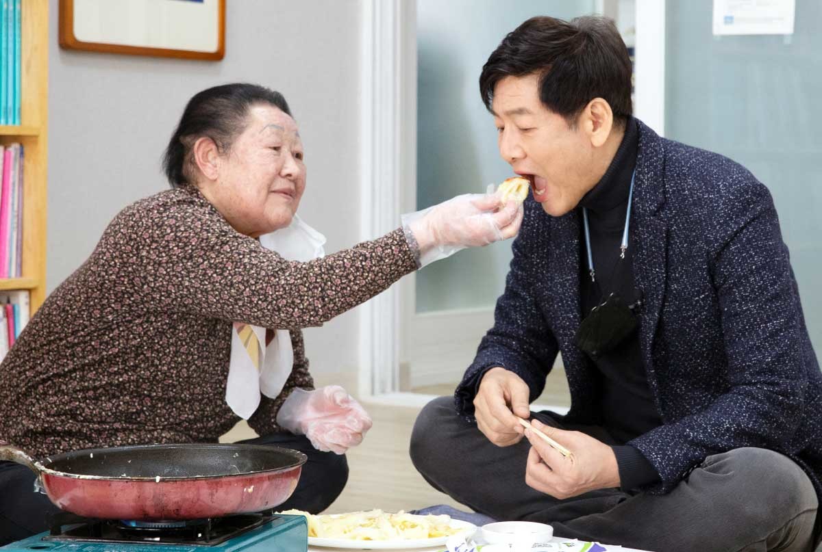 '칠곡할매글꼴' 홍보대사를 맡고 있는 방송인 정재환 씨가 지난 2021년 경북 칠곡군 금남2리 마을회관에서 글씨체를 만든 할머니가 만든 배추전을 받아 먹고 있다. 칠곡군은 지난 2020년 6월부터 성인문해교육을 통해 한글을 깨우친 할머니 400명 중 개성이 강한 글씨체를 선정해 5명의 글씨를 글꼴로 제작했다. / 칠곡군 제공