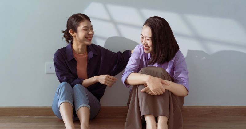 고요한 실내 공간에서 두 여인이 함께 즐거운 순간을 나누고 바닥에 앉아 창문을 통해 스치는 부드러운 햇살 아래 함께 웃고 있다. / 셔터스톡