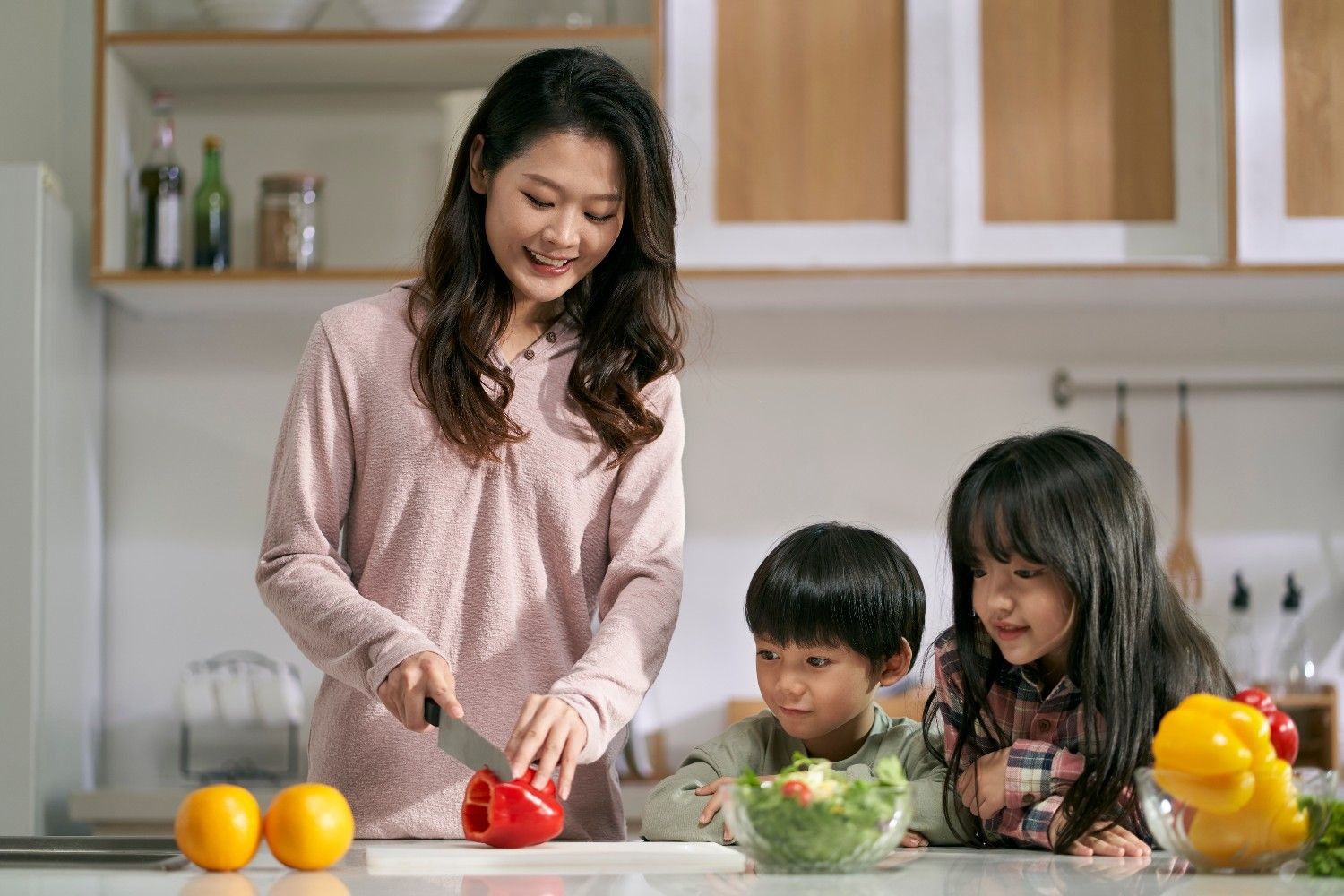 두 자녀가 옆에서 지켜보는 동안 샐러드를 만들기 위해 피망을 자르는 젊은 아시아 어머니 / 셔터스톡