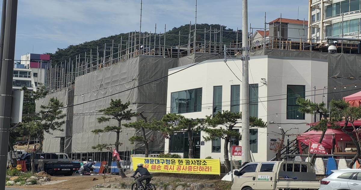 부산 해운대구 송정해수욕장 인근 광어골 해안가 건축 공사 현장에 대형 가설 구조물과 비계가 설치된 가운데, 주민들이 설치한 것으로 보이는 '해운대구청은 현 공사장 즉시 공사중단하라'는 내용의 현수막이 걸려 있다. 주민들은 건축허가 과정의 특혜 의혹과 해안 조망권 침해 등을 주장하며 반발하고 있다.