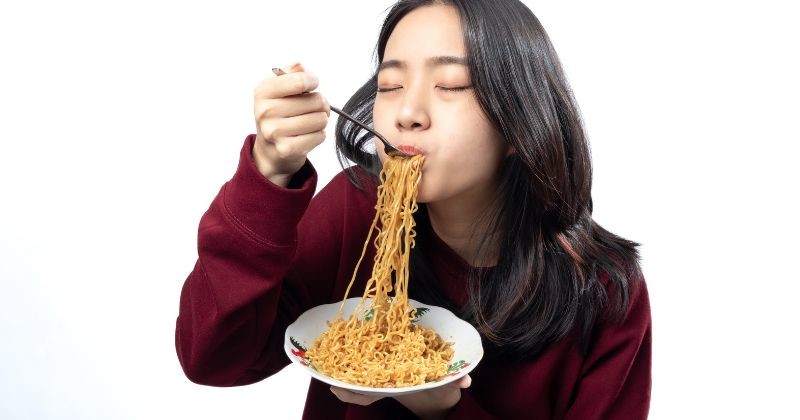 버건디 스웨터를 입고 하얀 배경에 고립된 채 그릇에 담긴 맛있는 국수를 즐기는 즐거운 아시아 여성. / 셔터스톡