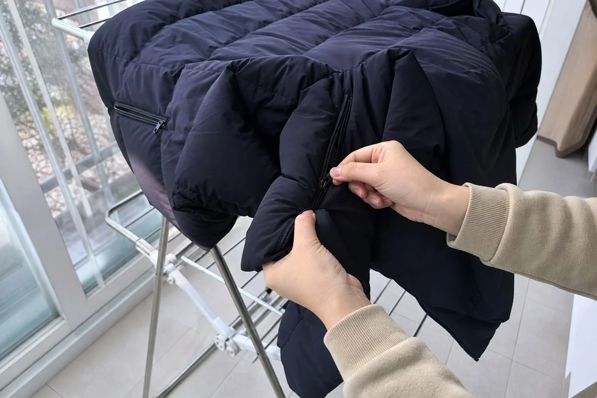 창가 빨래건조대에 걸린 검은 패딩 점퍼 지퍼를 손으로 여닫아 보는 모습 — 내용 이해를 돕는 AI 이미지