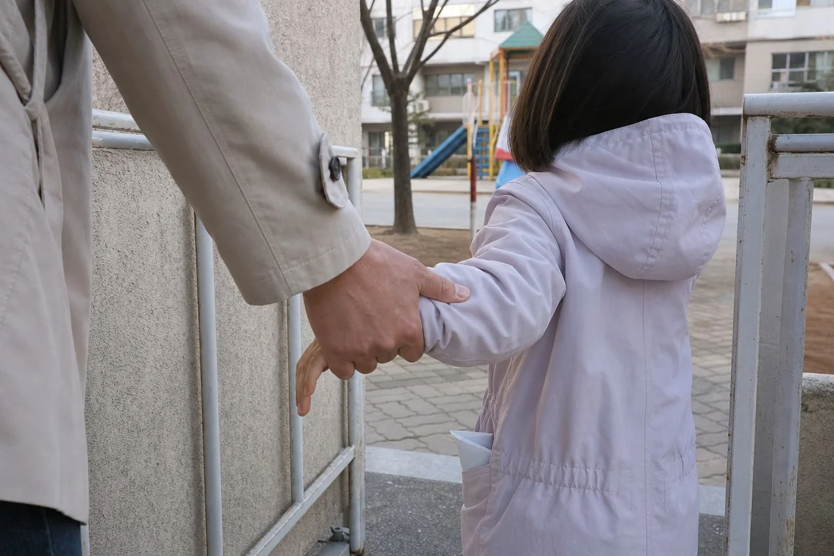 놀이터 철문 앞에서 연보라색 외투를 입은 어린이 손을 잡는 성인 — 이해를 돕는 AI 자료 이미지