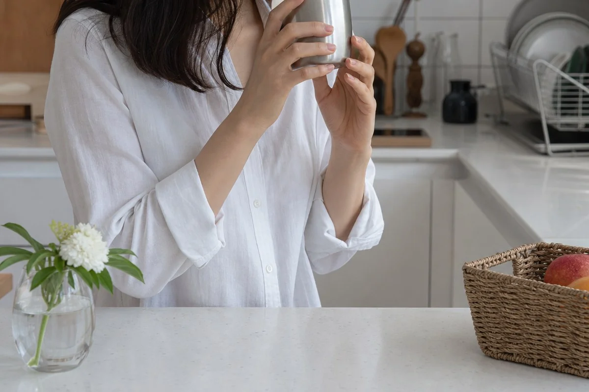 주방 식탁 앞에서 두 손으로 은색 컵을 입 가까이 들어 올린 모습 — 이해를 돕는 AI 자료 이미지