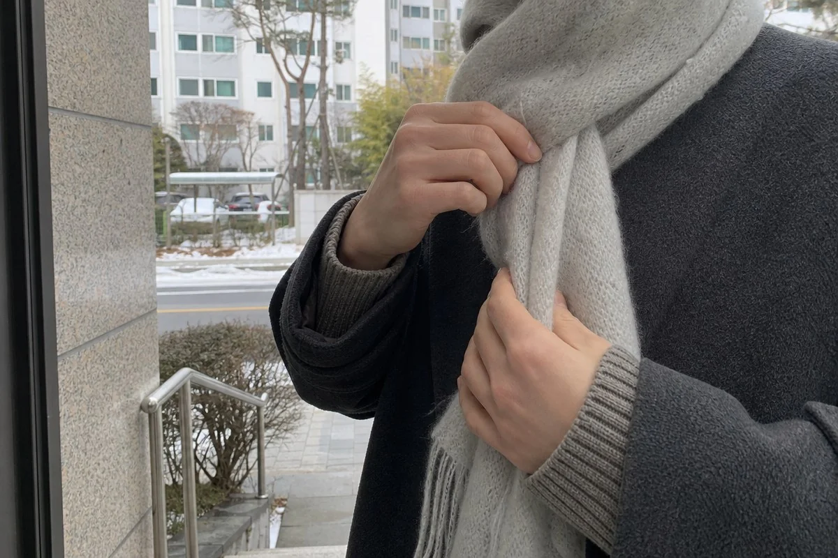 눈 쌓인 건물 앞에서 긴 목도리를 여미는 모습 — 내용 이해를 돕는 AI 이미지