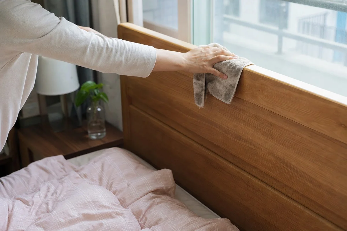 침실에서 침대 곁 창가의 나무 턱을 천으로 닦는 모습 — 이해를 돕는 AI 자료 이미지