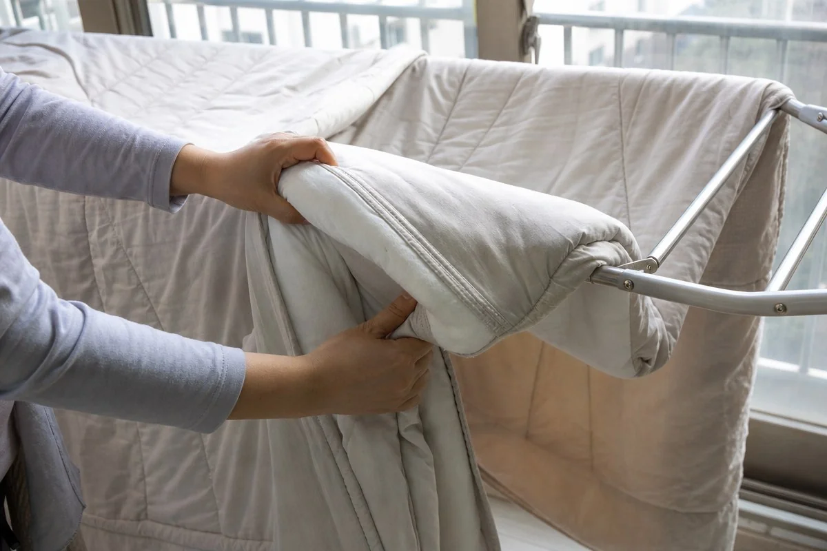 창가에서 두 손으로 이불을 빨래건조대에 거는 모습 — 내용 이해를 돕는 AI 이미지